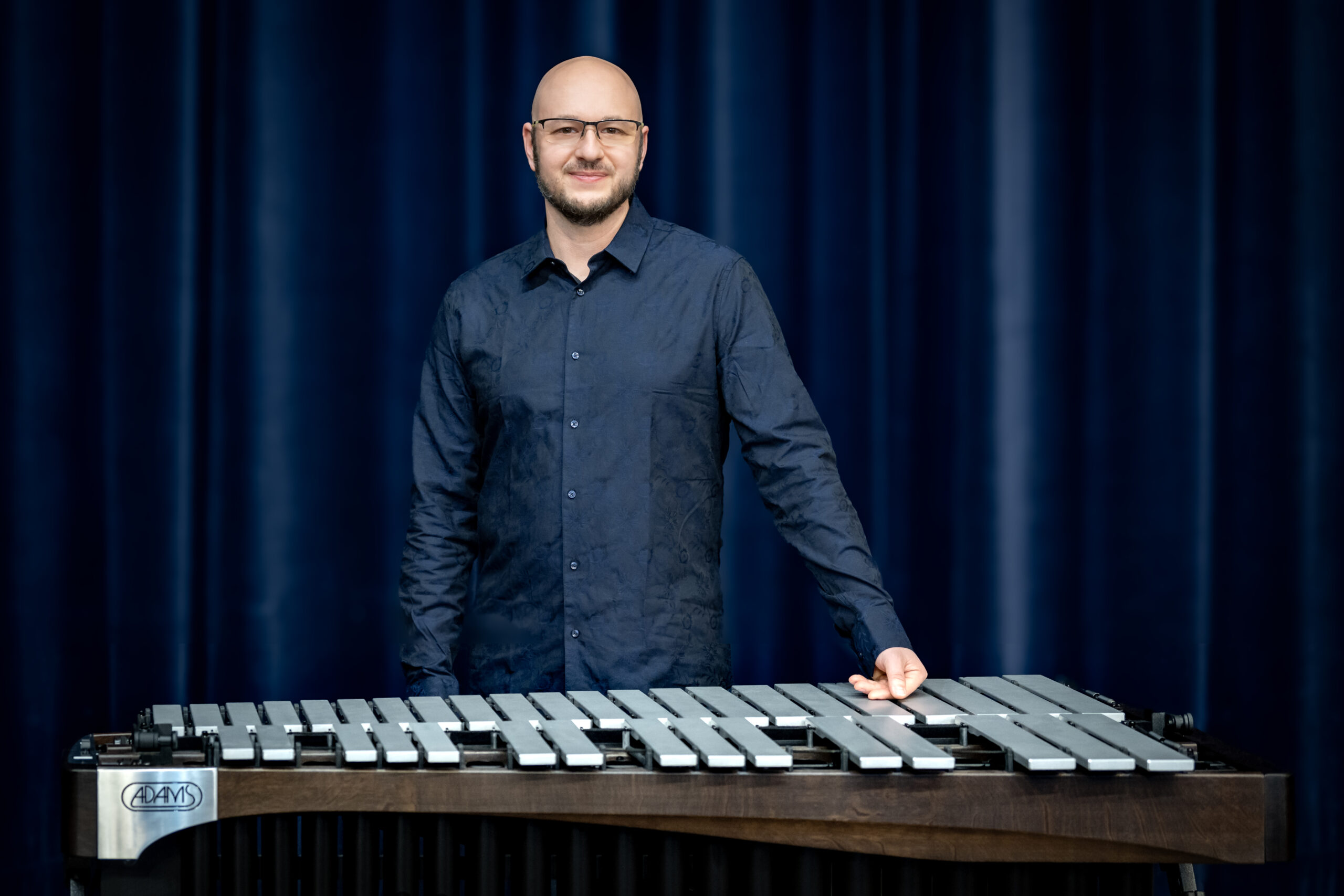 Wojciech Herzyk za instrumentem perkusyjnym na tle ciemnej kurtyny.