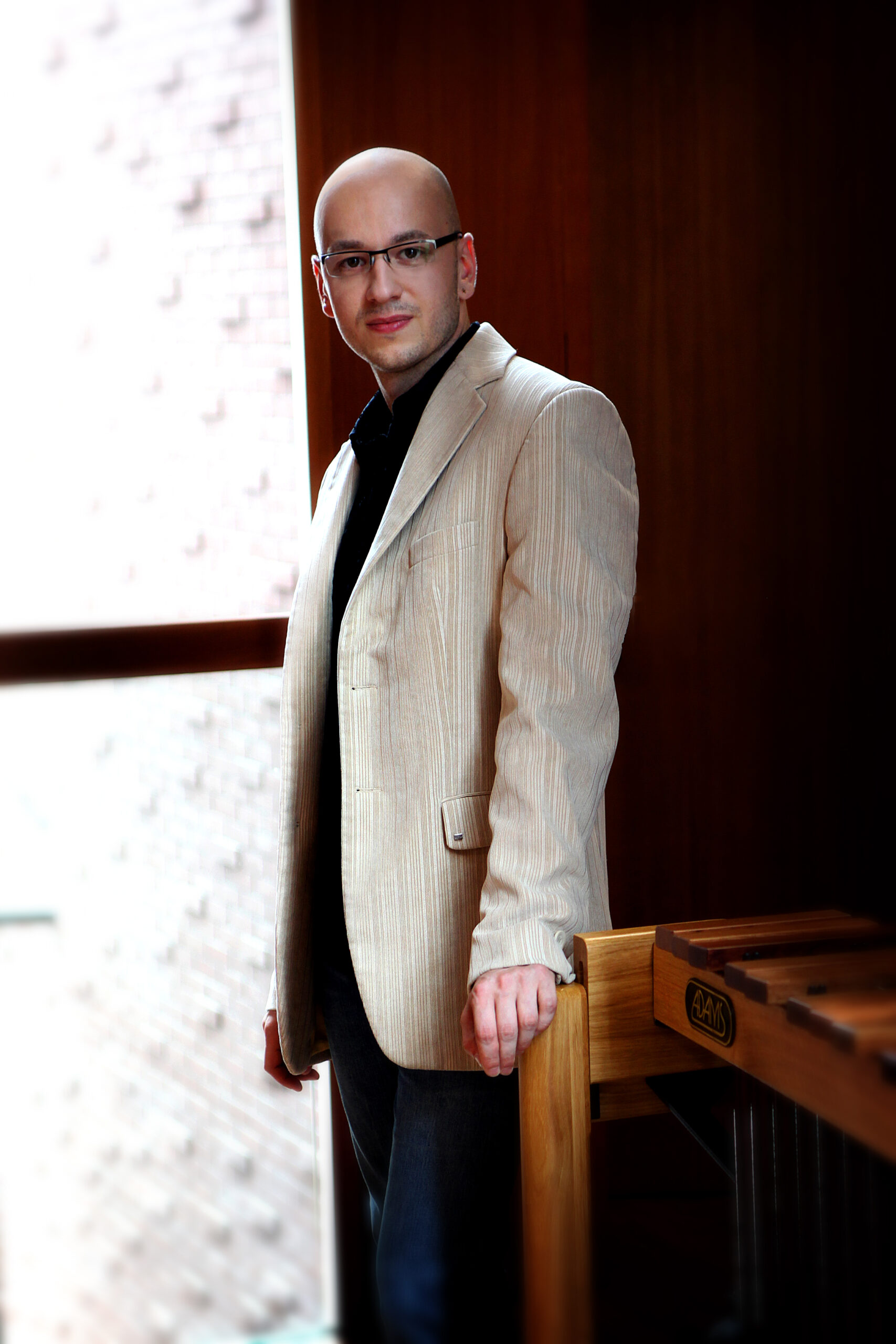 Wojciech Herzyk przy drewnianym instrumencie, ubrany w jasną marynarkę.